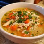 A close-up of a bowl of hearty chicken and rice soup, filled with shredded chicken, carrots, celery, rice, and parsley.
