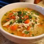 A close-up of a bowl of hearty chicken and rice soup, filled with shredded chicken, carrots, celery, rice, and parsley.