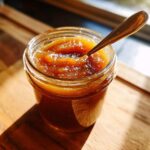 Close-up of a glass jar filled with rich, dark apple butter, a spoon resting inside.