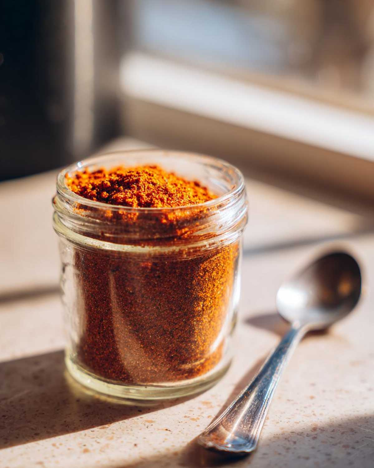 A small glass jar filled with homemade taco seasoning, next to a silver spoon.