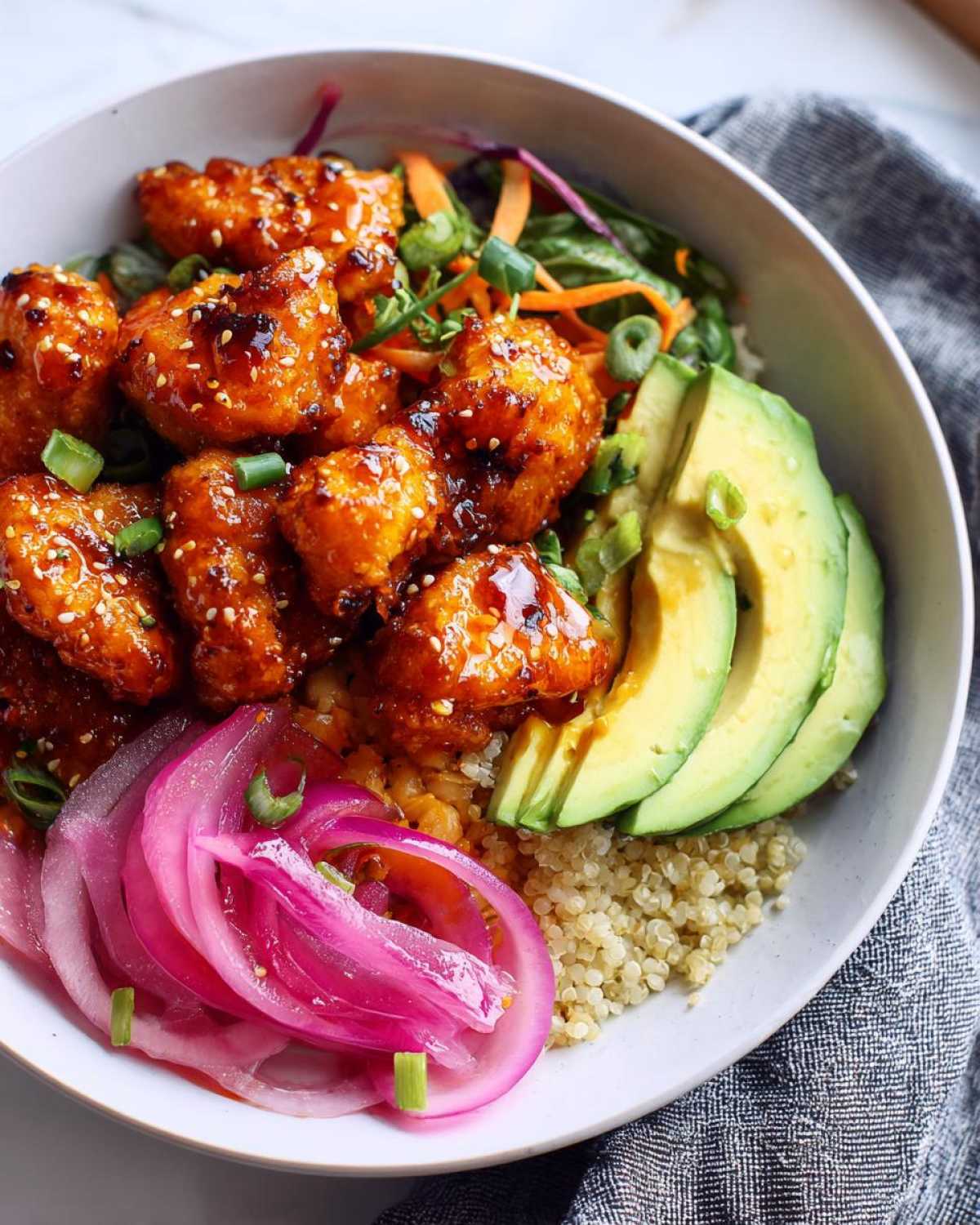 A close-up of a delicious hot honey chicken bowl, featuring glazed chicken, quinoa, sliced avocado, and pickled red onions.