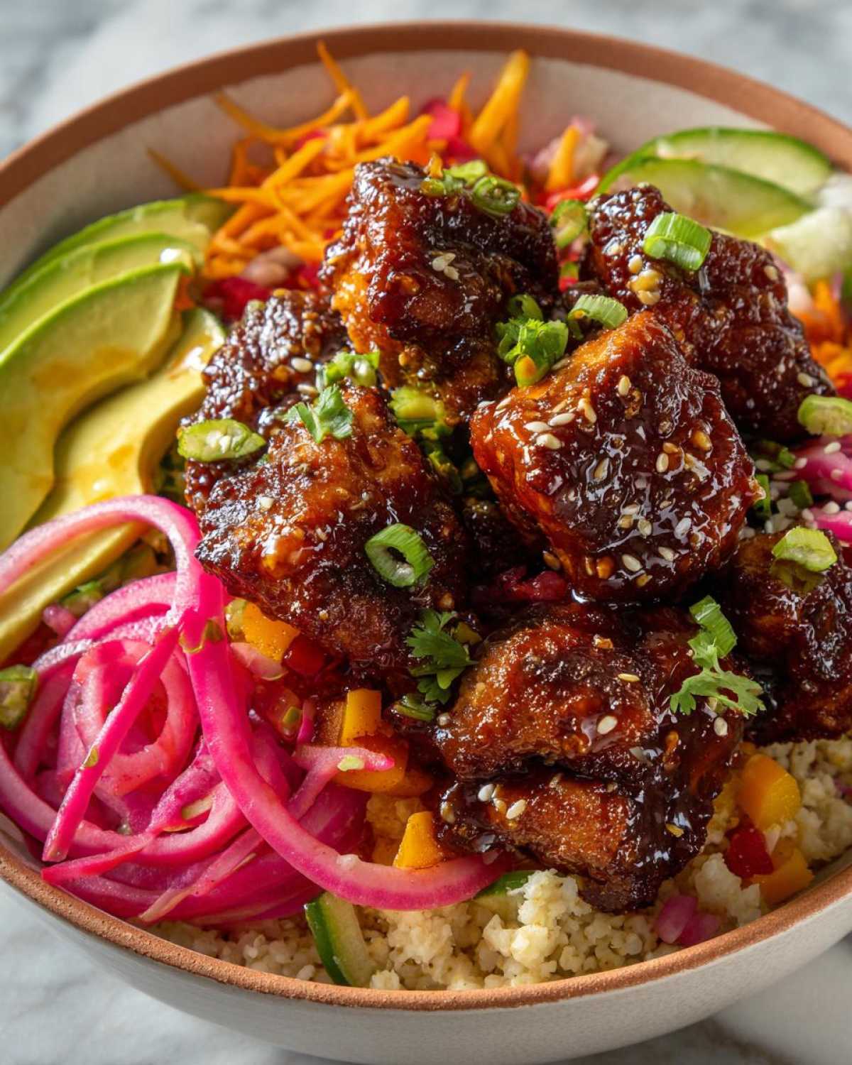 A close-up of a delicious Hot Honey Chicken Bowl featuring glazed chicken pieces, avocado, pickled onions, shredded carrots, and cucumber.