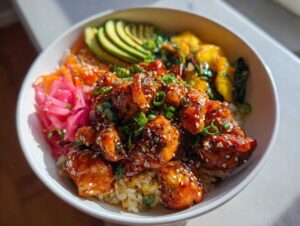 A close-up of a vibrant Hot Honey Chicken Bowl, featuring glazed chicken, rice, sliced avocado, pickled red onions, and greens.
