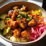 A close-up of a hot honey chicken bowl with glazed chicken, avocado, pickled onions, and shredded vegetables on rice.