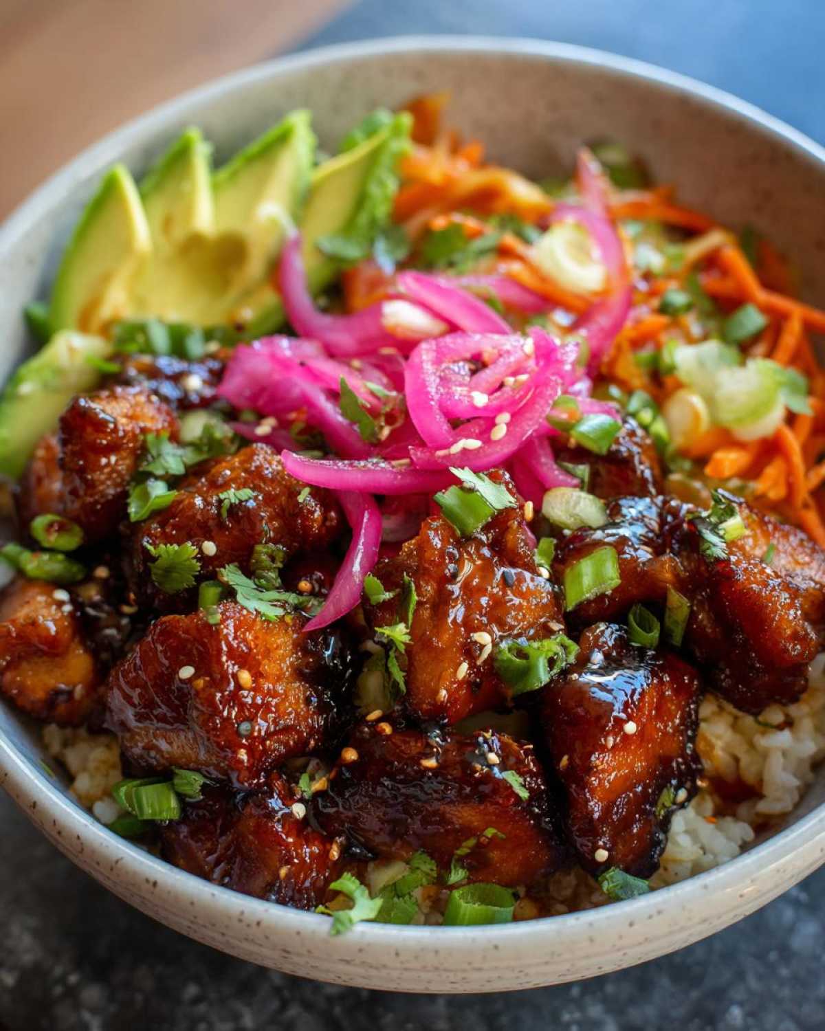 A close-up of a delicious Hot Honey Chicken Bowl, featuring glazed chicken pieces, sliced avocado, pickled red onions, and shredded carrots.