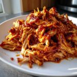 A close-up of a white plate piled high with tender, shredded Instant Pot pulled BBQ chicken coated in a rich, glossy BBQ sauce.