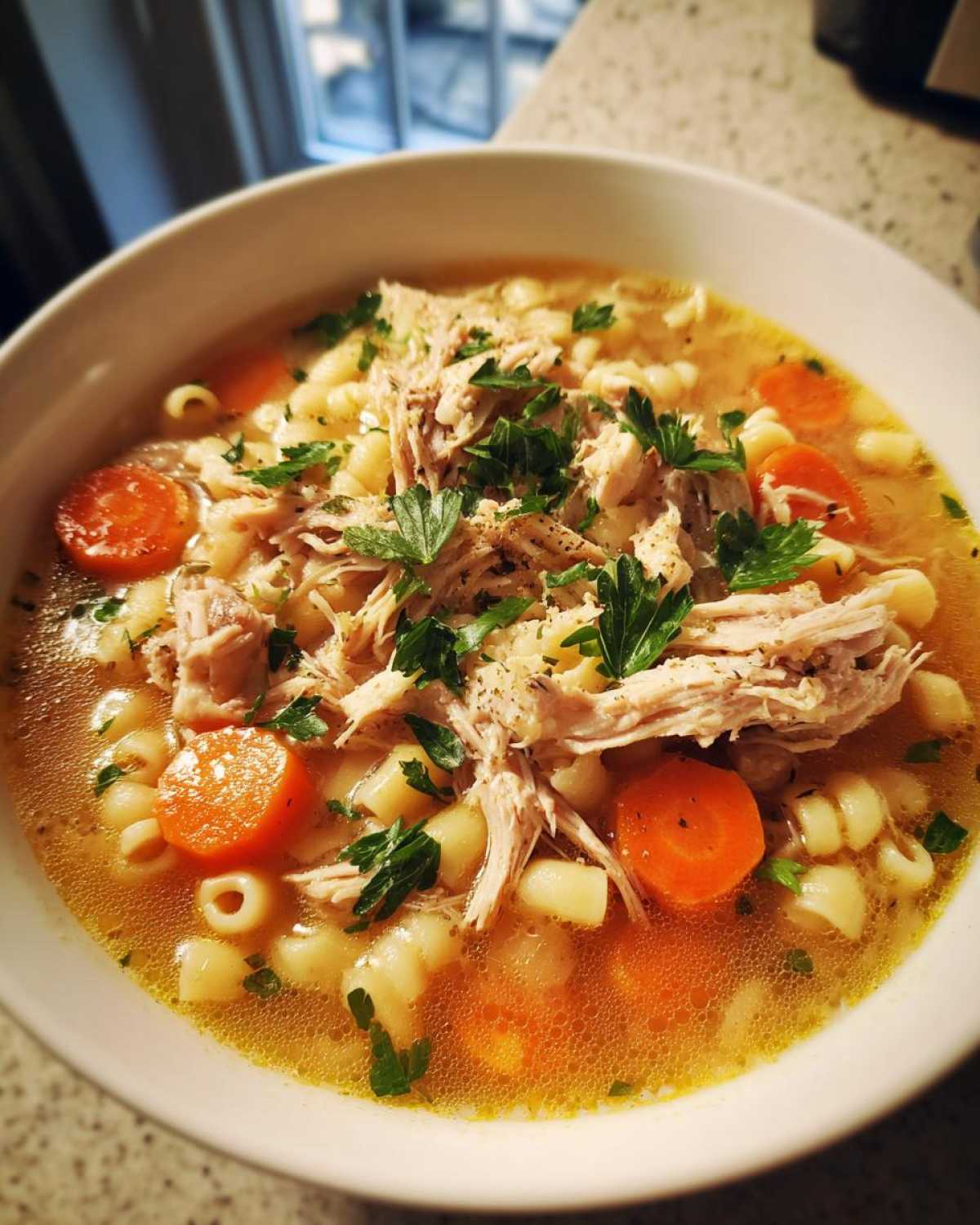A close-up of a bowl of Italian penicillin soup, featuring shredded chicken, ditalini pasta, and sliced carrots, garnished with fresh parsley.