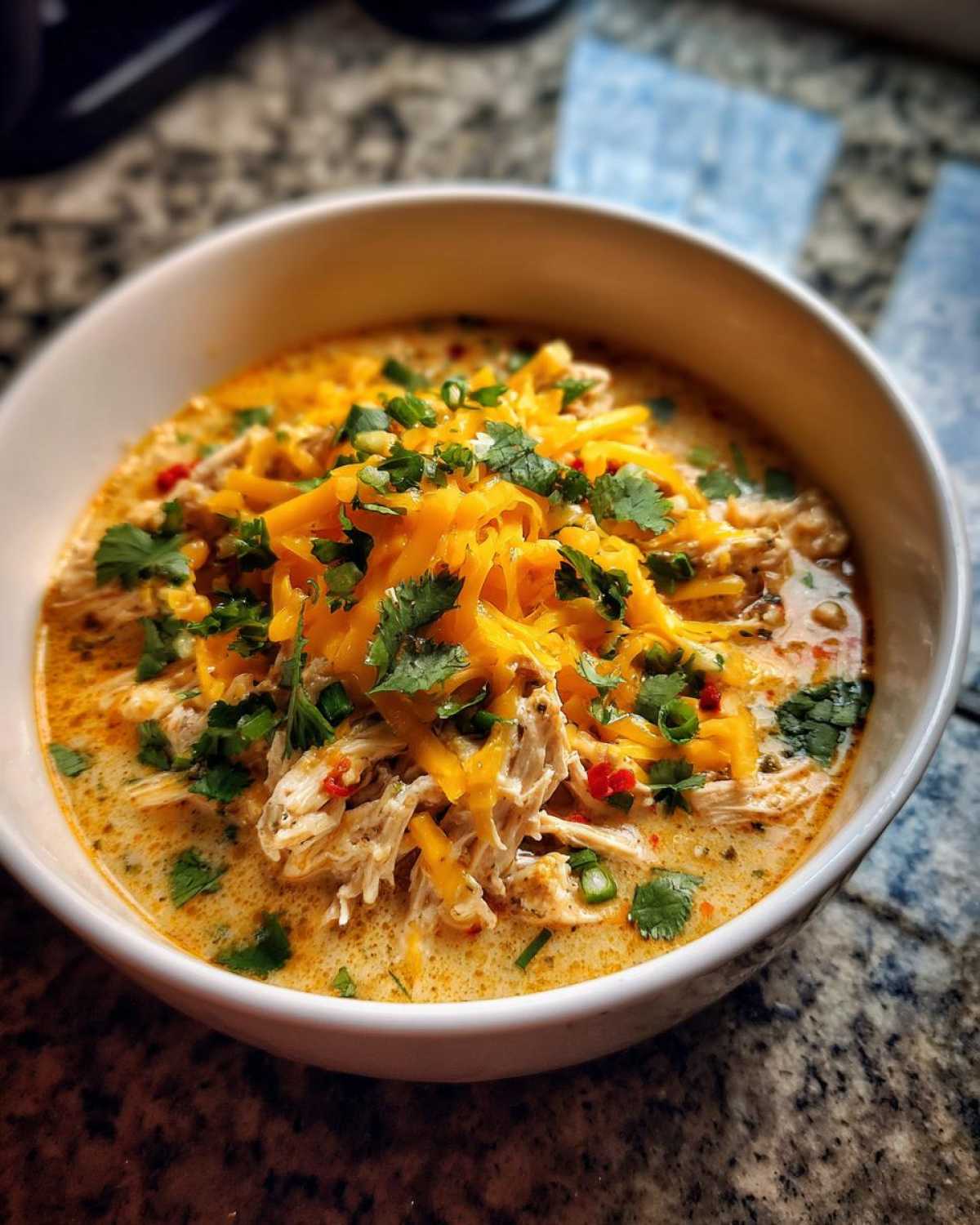A close-up of a white bowl filled with creamy Keto Cream Cheese Chicken Chili, topped with shredded cheddar cheese, cilantro, and green onions.