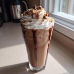 A tall glass filled with a frothy Kinder Chocolate Frappuccino, topped with whipped cream, chocolate chunks, and cookie crumbles.