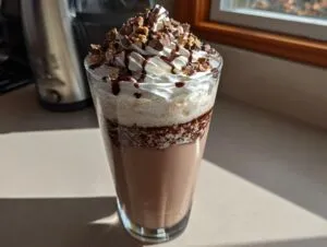 A tall glass filled with a creamy Kinder Chocolate Frappuccino, topped with whipped cream, chocolate drizzle, and chopped candy.