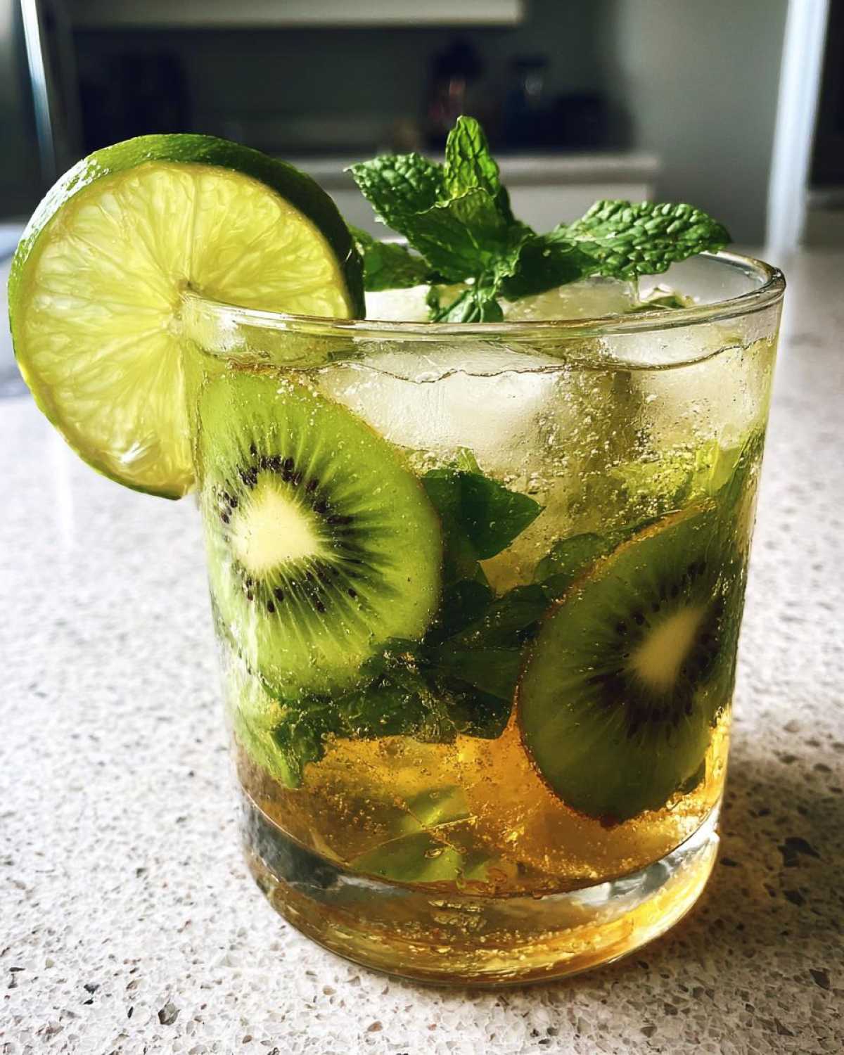 A close-up of a refreshing Kiwi Mojito cocktail, featuring slices of kiwi, fresh mint, a lime wedge, and ice.