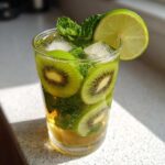 A tall glass filled with a refreshing Kiwi Mojito, featuring slices of kiwi, fresh mint, ice, and a lime wedge.