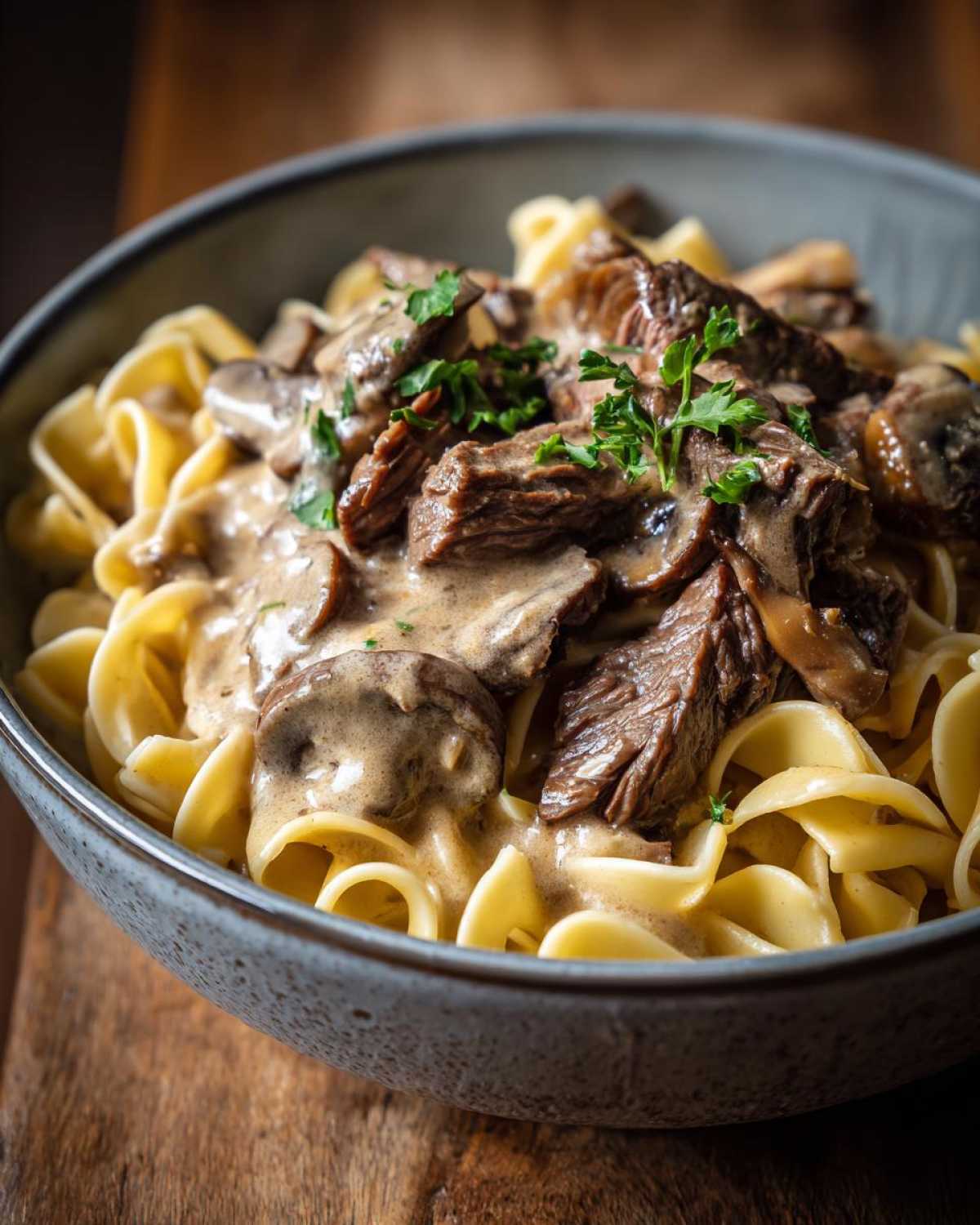 A close-up of a bowl of old fashioned beef stroganoff served over wide egg noodles, garnished with parsley.