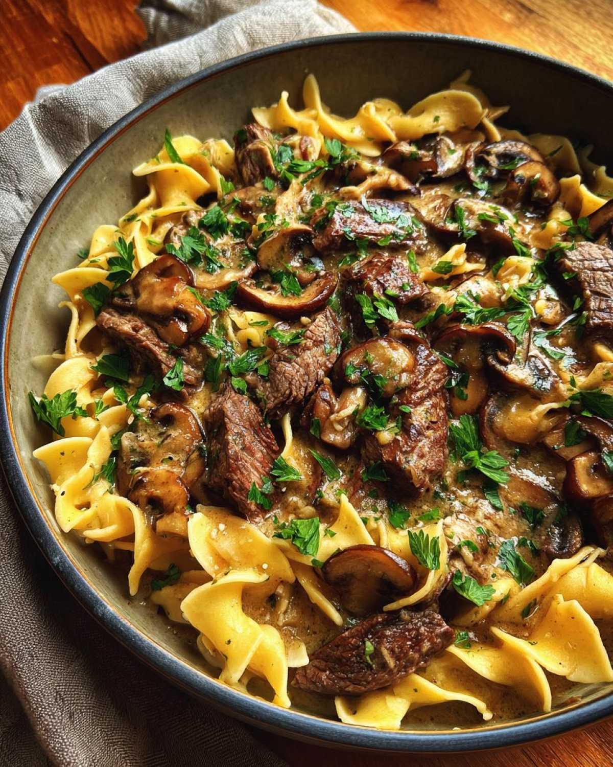 A close-up of a bowl of old fashioned beef stroganoff served over wide egg noodles, topped with mushrooms and parsley.
