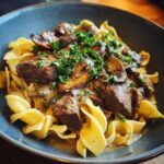 A close-up of a bowl of old fashioned beef stroganoff served over egg noodles, topped with mushrooms and parsley.