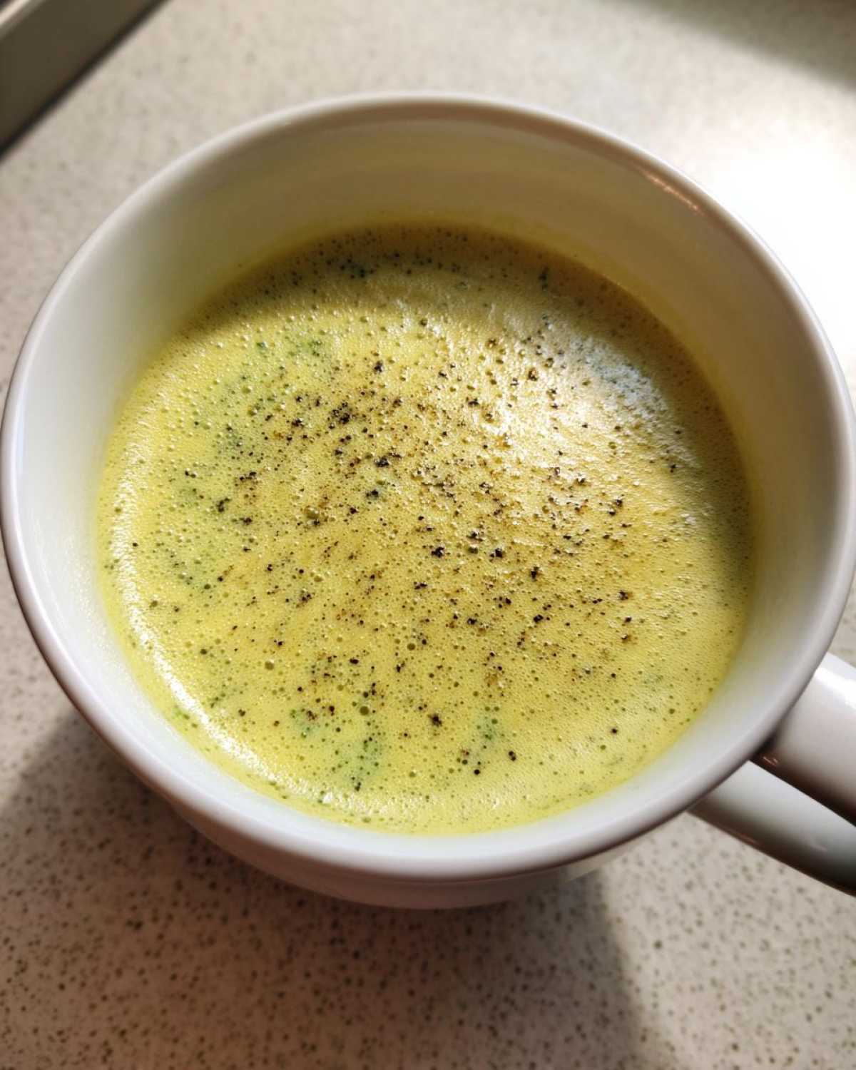 Close-up of a steaming mug of Panera Broccoli Cheddar Soup, topped with black pepper.