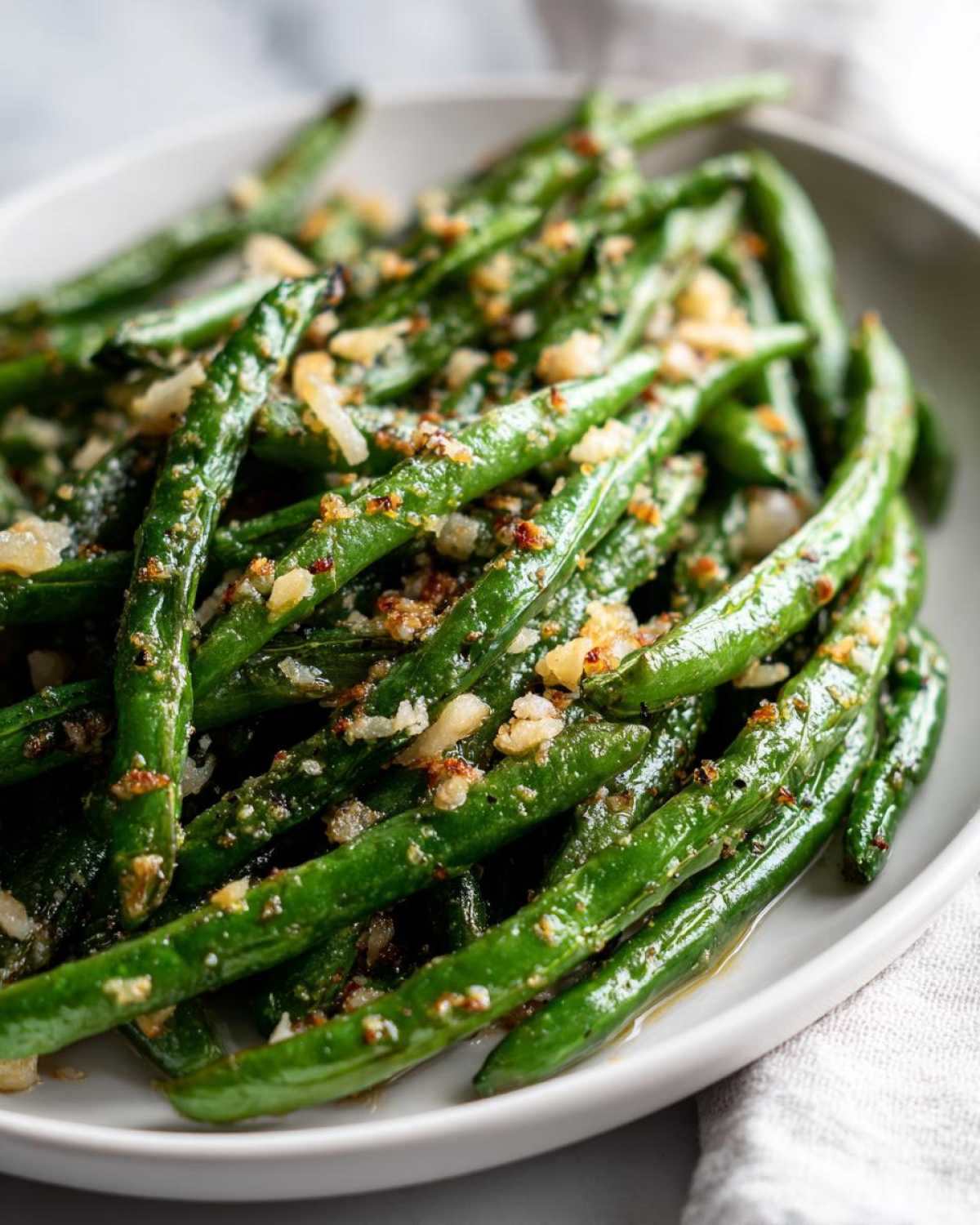 Close-up of a white bowl filled with glossy, roasted parmesan green beans, topped with crispy garlic and parmesan.