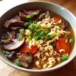 A steaming bowl of slow cooker beef ramen noodles, featuring tender beef slices, ramen noodles, mushrooms, carrots, and green onions.