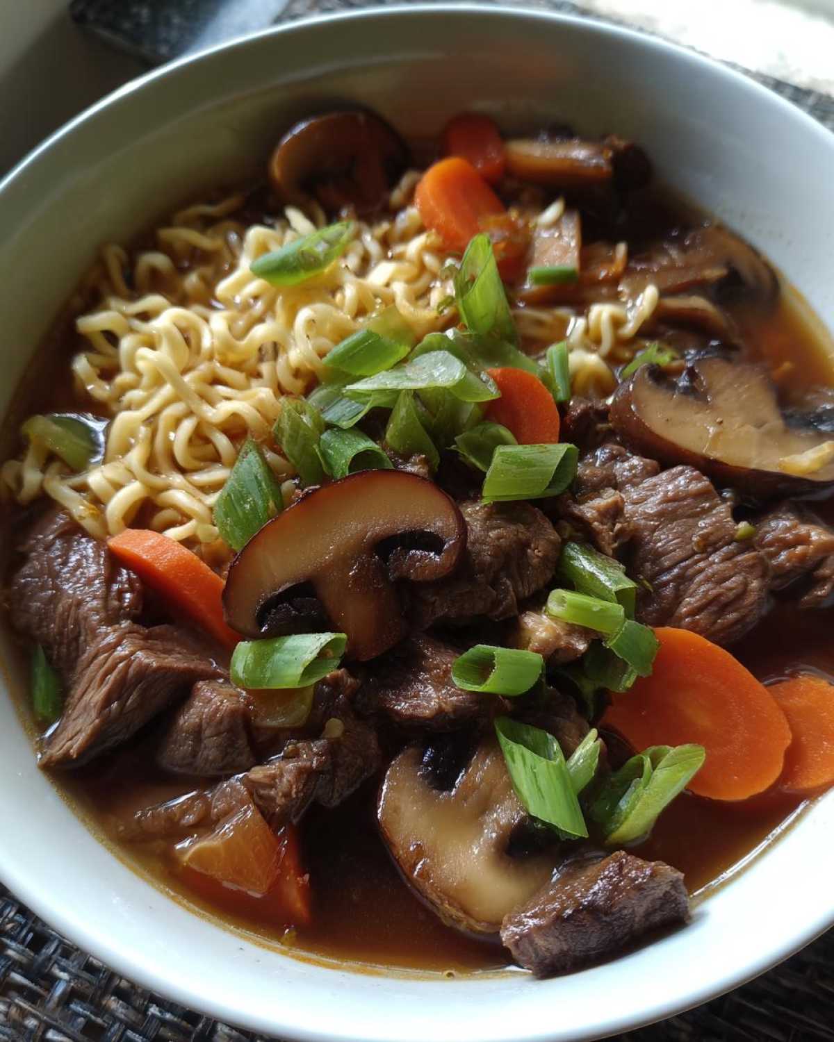 A close-up of a bowl of slow cooker beef ramen noodles, featuring tender beef, mushrooms, carrots, and green onions.