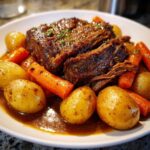 A close-up of a tender slow cooker pot roast, sliced and served with baby potatoes and carrots in a rich gravy.