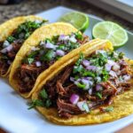 Three delicious slow cooker shredded beef tacos topped with red onion and cilantro, served with lime wedges.
