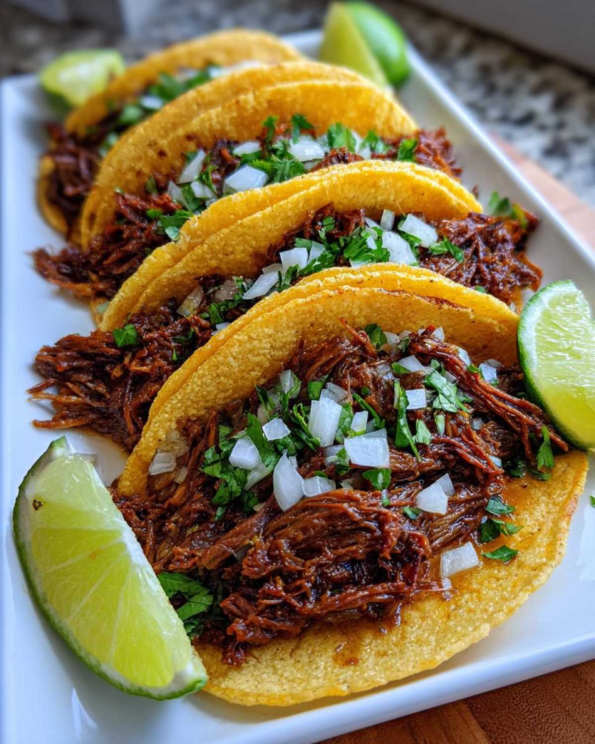 A plate of four delicious slow cooker shredded beef tacos, topped with diced onions and cilantro, served with lime wedges.