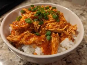 A bowl of fluffy white rice topped with shredded slow cooker sweet chili chicken and garnished with chopped green onions.
