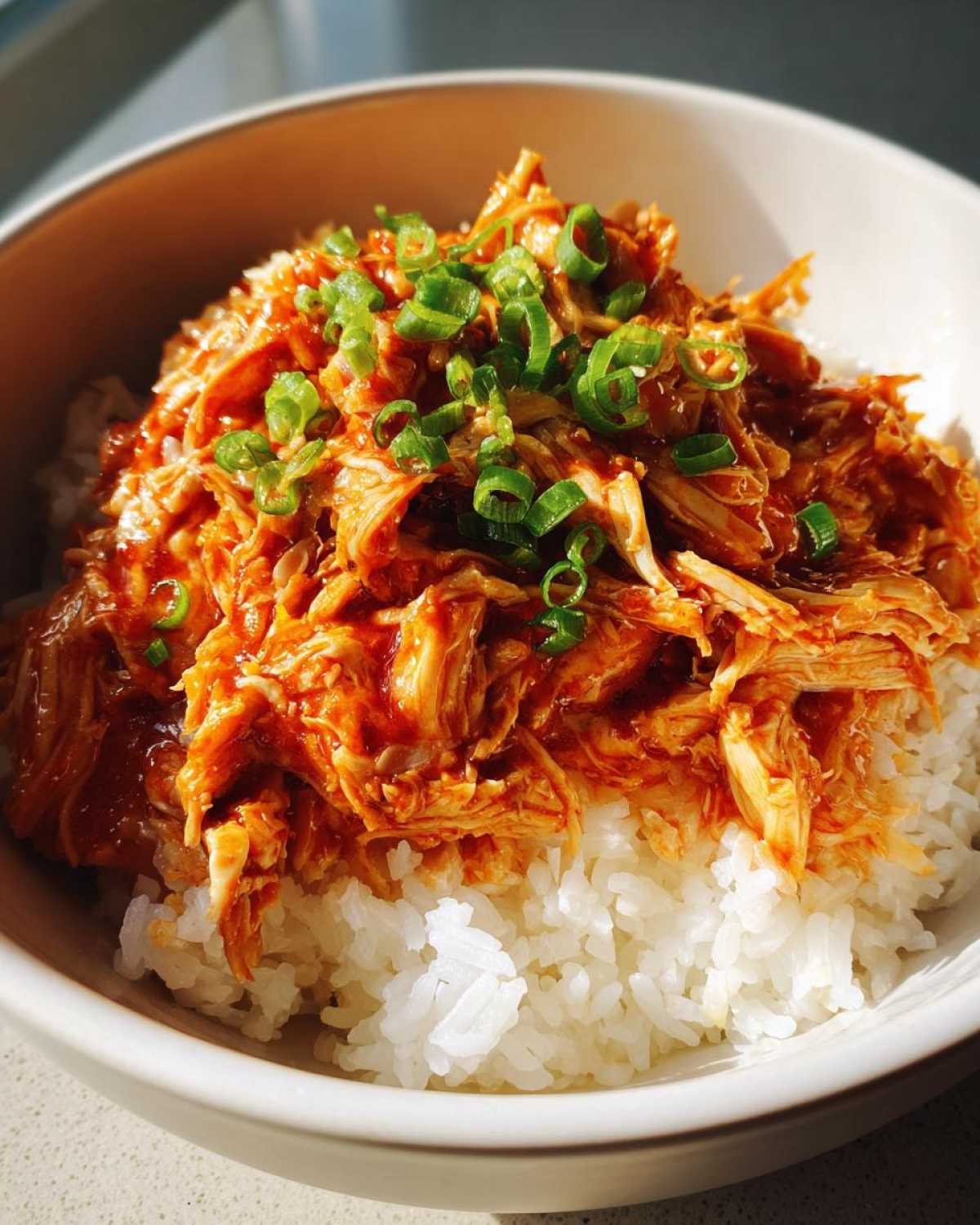 A bowl of white rice topped with shredded slow cooker sweet chili chicken and garnished with chopped green onions.