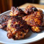 A close-up of several crispy southern baked chicken thighs on a white plate, with a dark, seasoned crust.