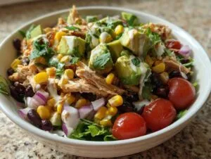 A vibrant bowl of Southwest chicken salad featuring shredded chicken, black beans, corn, avocado, red onion, and tomatoes, drizzled with dressing.