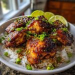 A bowl of fluffy white coconut rice topped with glistening, glazed spicy maple chicken pieces and garnished with fresh scallions and lime wedges.