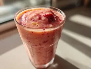 A close-up of a tall glass filled with a vibrant pink Strawberry Mango Smoothie, with visible fruit pieces.
