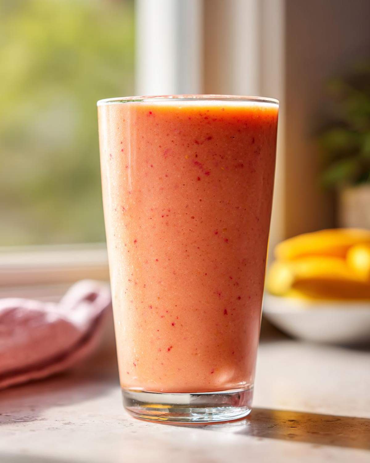 A tall glass filled with a vibrant pink Strawberry Mango Smoothie, with tiny red flecks visible.