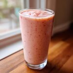A tall glass filled with a creamy pink Strawberry Pina Colada Smoothie, showing small red strawberry pieces throughout.