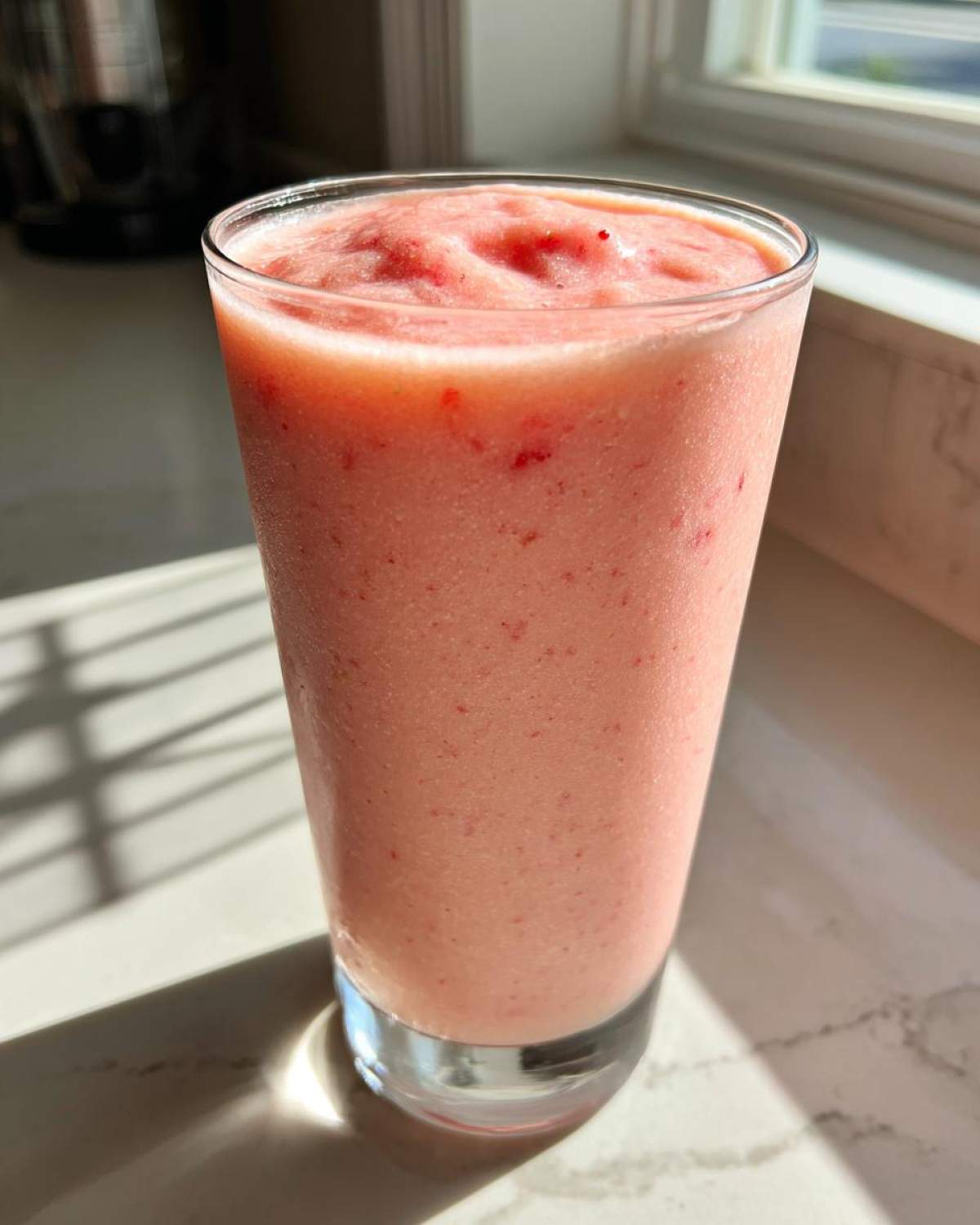 A tall glass filled with a vibrant pink Strawberry Pina Colada Smoothie, showing flecks of strawberry.