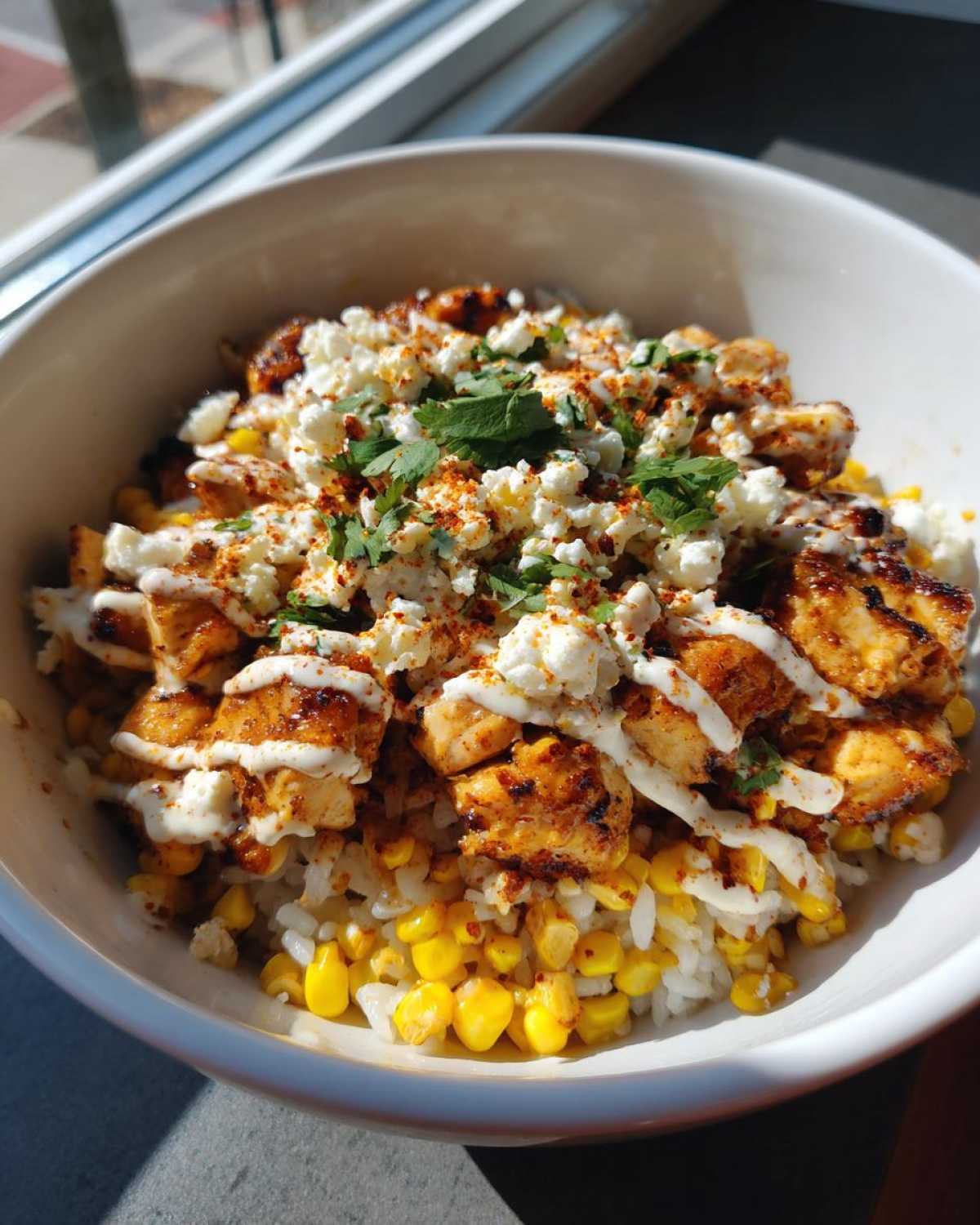 A close-up of a street corn chicken and rice bowl, featuring grilled chicken, corn, rice, crumbled cheese, and cilantro.