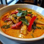 Close-up of a bowl of creamy Thai red curry chicken with red bell peppers and basil leaves.