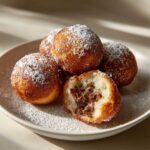 A plate of golden brown air fryer Nutella bombs, dusted with powdered sugar. One is broken open to reveal a gooey Nutella filling.