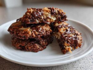 A stack of four rich, chewy almond joy cookies drizzled with melted dark chocolate on a white plate.