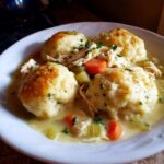 Four fluffy, golden-brown dumplings served over shredded chicken and vegetables in a creamy sauce, a perfect bowl of chicken and dumplings.