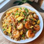 A close-up of a white bowl filled with flavorful shrimp fried rice, topped with large shrimp, scrambled egg pieces, and chopped green onions.