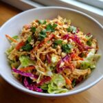 A vibrant bowl of thai chicken salad featuring shredded chicken, cabbage, carrots, and topped with crushed peanuts and cilantro.