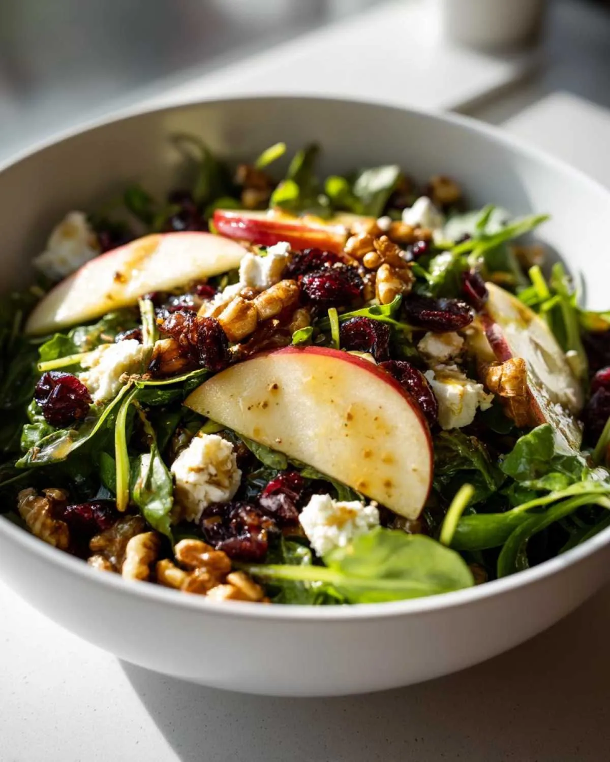 A close-up of a vibrant apple feta salad in a white bowl, featuring fresh greens, sliced apples, crumbled feta, walnuts, and dried cranberries.