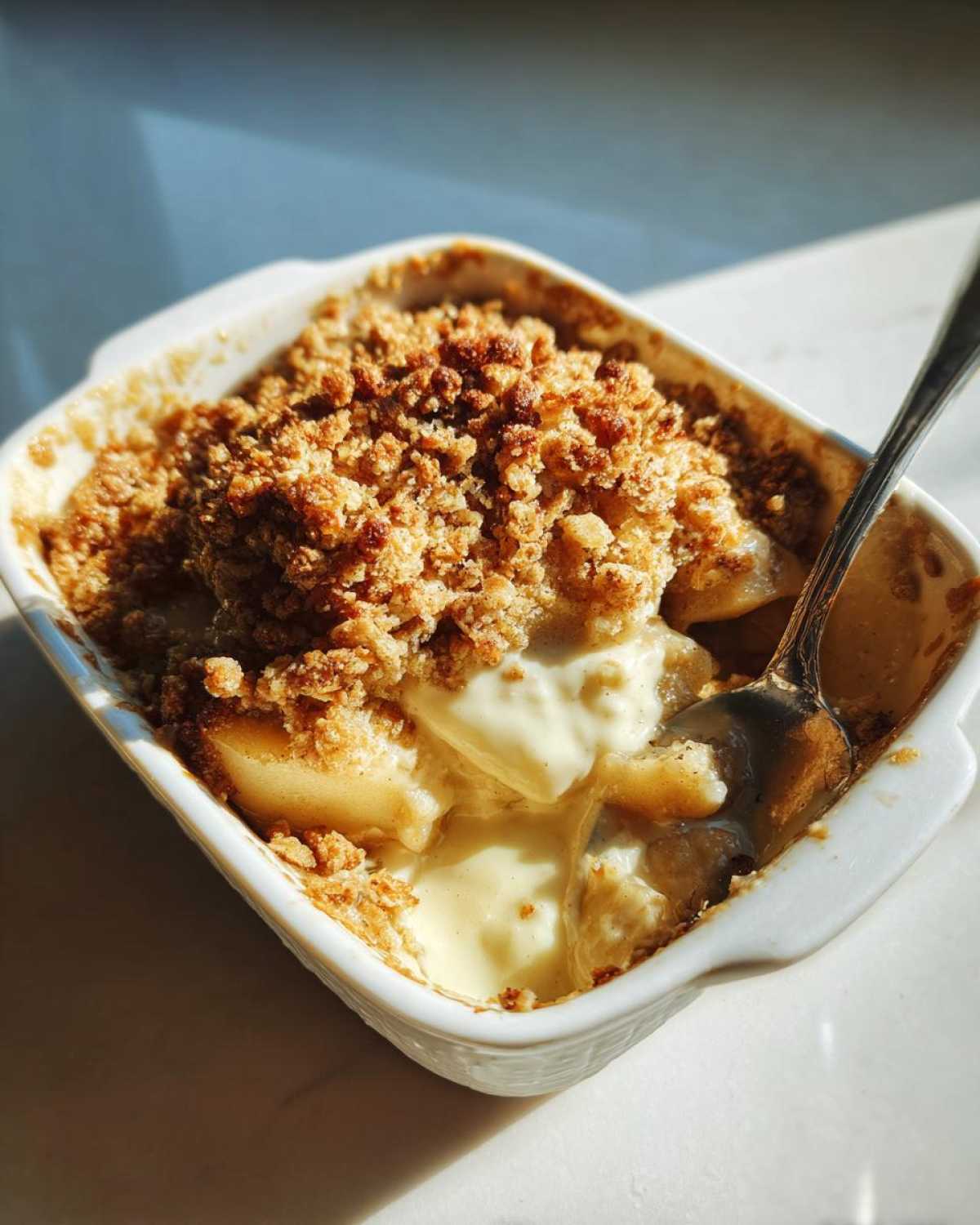 A close-up of warm Apple Pie Dip in a white baking dish, with a spoon scooping into the creamy, cinnamony apple filling and crumb topping.