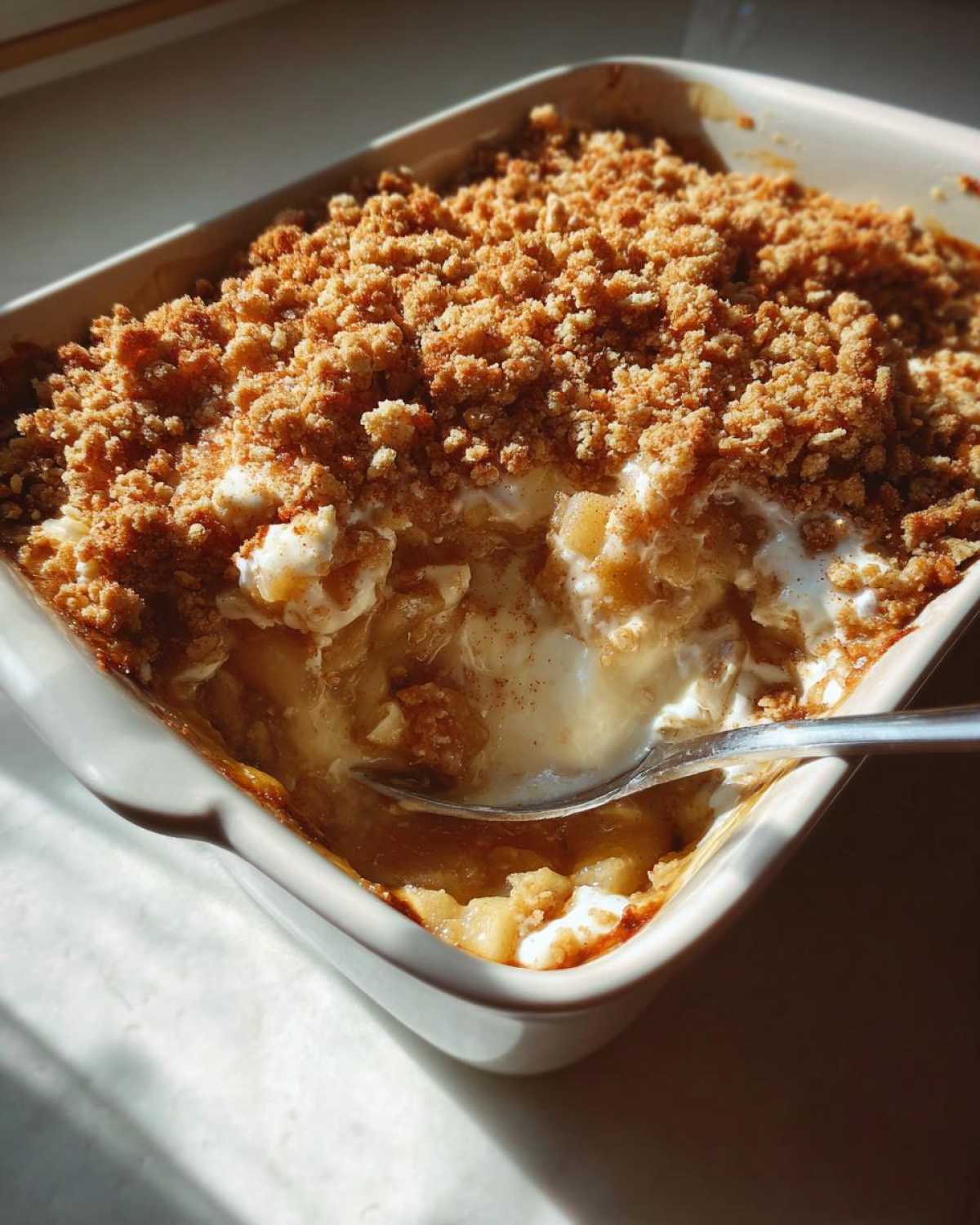 A close-up of a white baking dish filled with warm Apple Pie Dip, featuring a spoon digging into the gooey apple filling and crumb topping.
