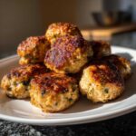 A close-up of a pile of golden-brown baked chicken ricotta meatballs on a white plate, sprinkled with herbs.
