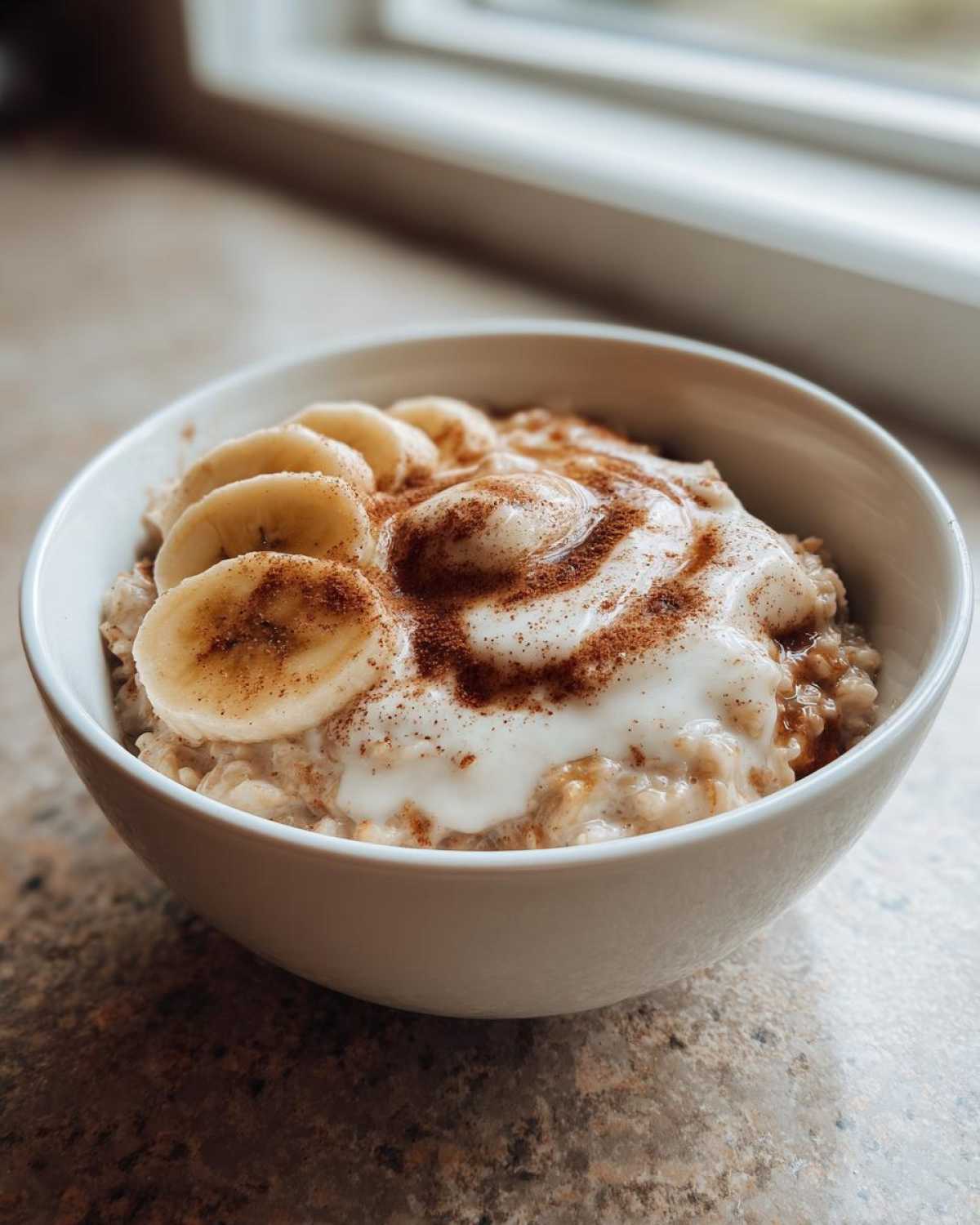 A close-up of a white bowl filled with creamy banana oatmeal, topped with sliced bananas, yogurt, and a dusting of cinnamon.
