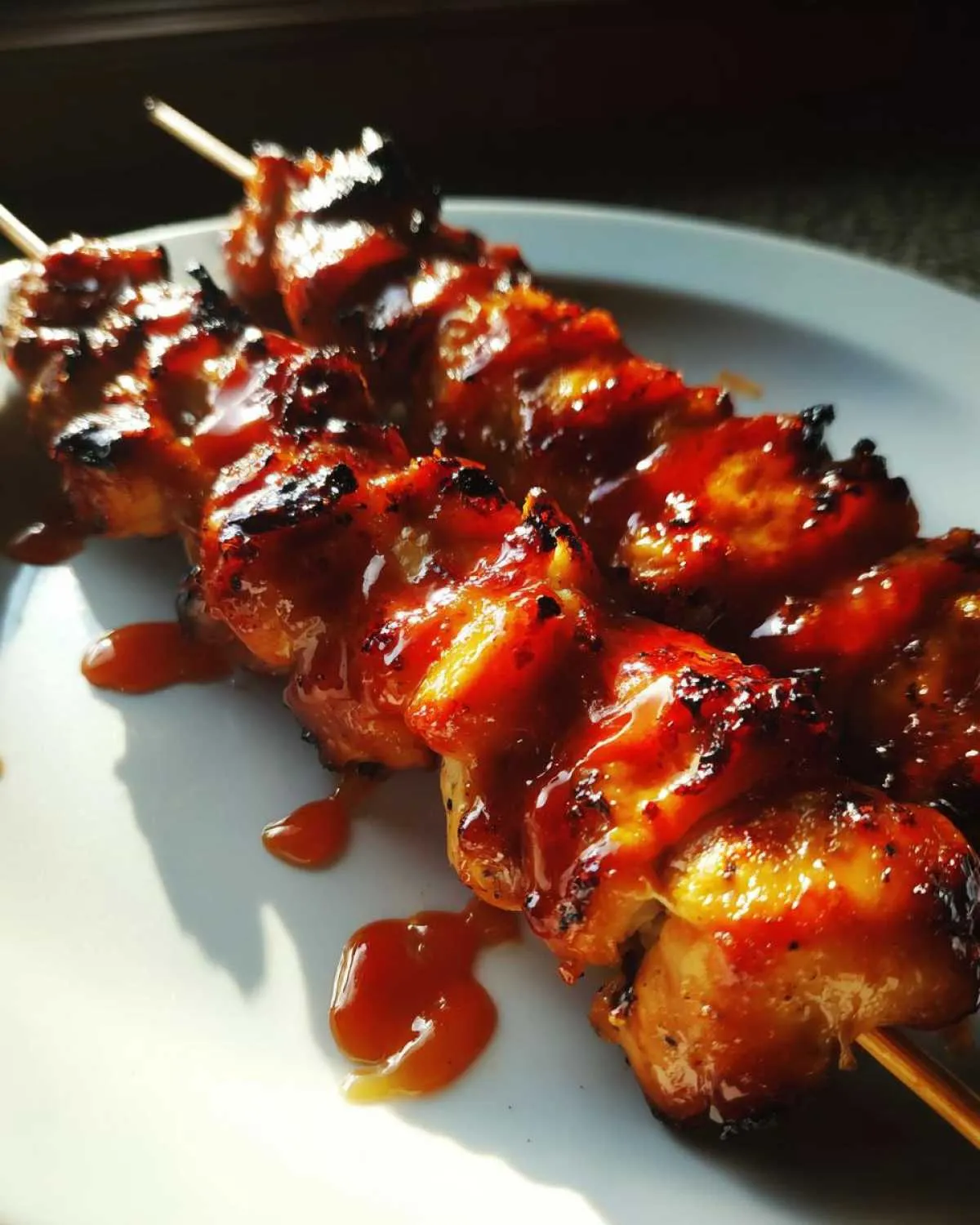Close-up of two juicy Bang Bang chicken skewers on a white plate, glazed with a rich, red sauce.