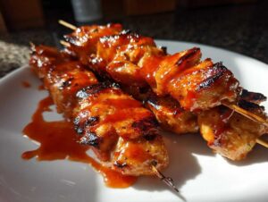 Three juicy Bang Bang chicken skewers coated in a glossy, orange sauce on a white plate.