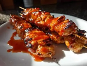 Three juicy Bang Bang chicken skewers coated in a glossy, orange sauce on a white plate.
